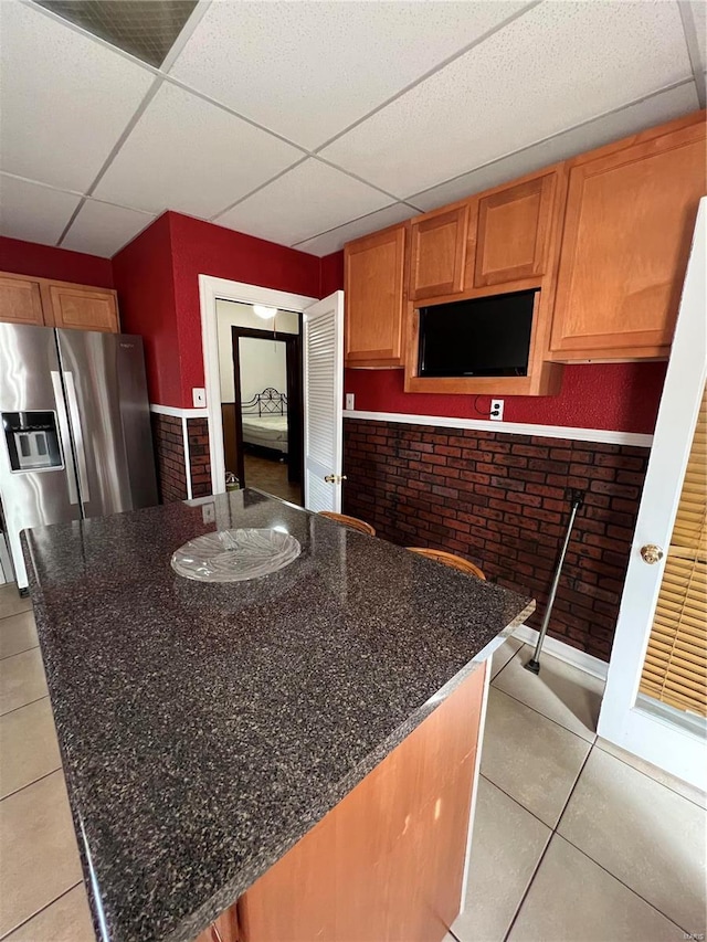 kitchen featuring a drop ceiling, brick wall, dark stone countertops, stainless steel fridge, and light tile patterned flooring