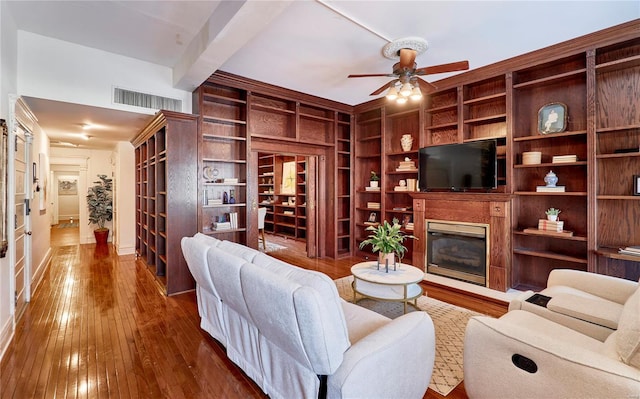 living room featuring a ceiling fan, visible vents, dark wood-style flooring, a glass covered fireplace, and beamed ceiling