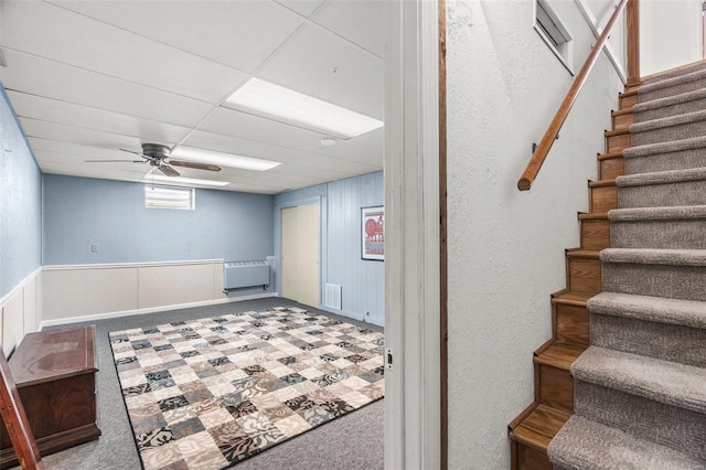 basement featuring carpet, stairway, radiator heating unit, ceiling fan, and a drop ceiling