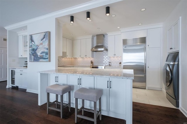 kitchen with wall chimney range hood, washer / dryer, white cabinetry, light stone counters, and built in fridge