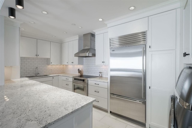 kitchen featuring white cabinetry, appliances with stainless steel finishes, wall chimney range hood, light stone countertops, and sink