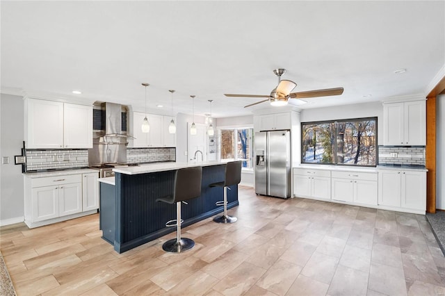 kitchen with a center island with sink, stainless steel fridge with ice dispenser, wall chimney exhaust hood, white cabinetry, and decorative light fixtures