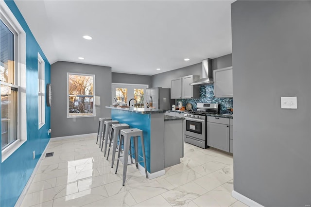 kitchen with gray cabinets, a center island with sink, a breakfast bar, appliances with stainless steel finishes, and wall chimney exhaust hood