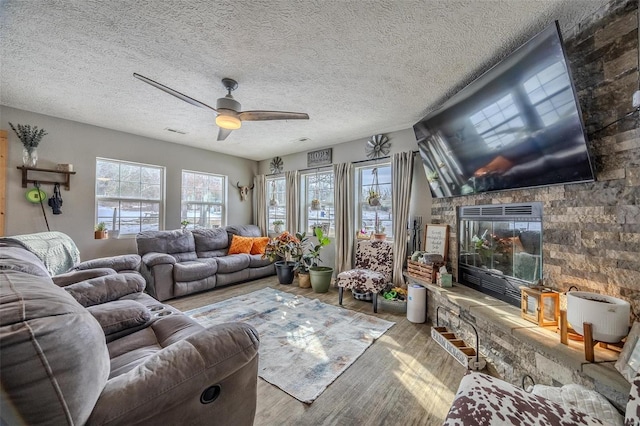 living room featuring ceiling fan, light wood-type flooring, a stone fireplace, and a textured ceiling