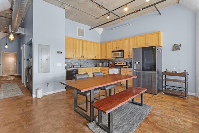 kitchen featuring electric panel, a high ceiling, stainless steel appliances, concrete flooring, and light brown cabinetry