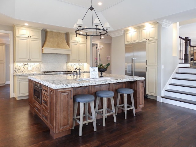 kitchen featuring built in appliances, a kitchen island with sink, light stone countertops, cream cabinets, and wall chimney range hood