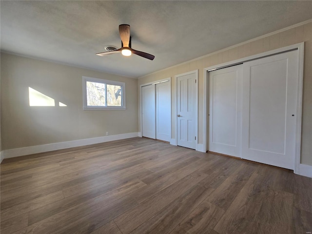 unfurnished bedroom featuring wood finished floors, a ceiling fan, baseboards, two closets, and crown molding