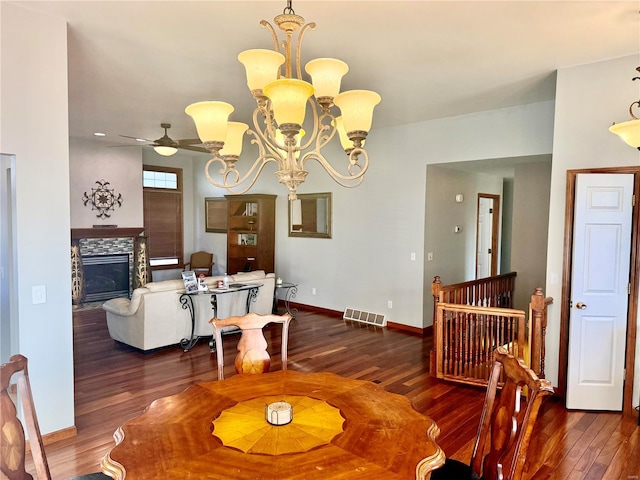 dining space with a stone fireplace, dark wood-type flooring, and ceiling fan with notable chandelier