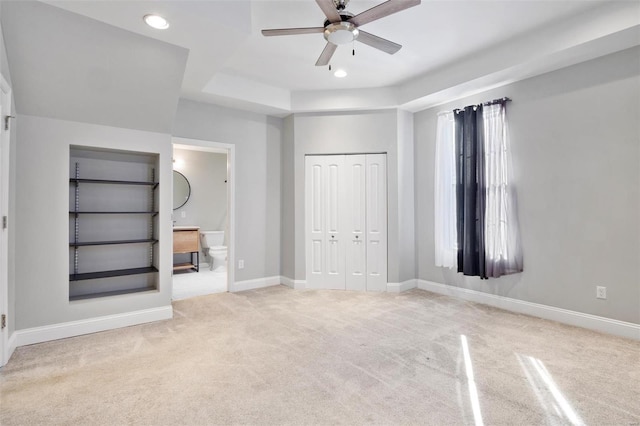 unfurnished bedroom with ensuite bath, light carpet, a tray ceiling, a closet, and ceiling fan