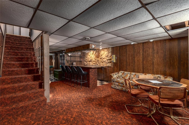 carpeted dining area featuring a dry bar, wooden walls, stairs, and a paneled ceiling