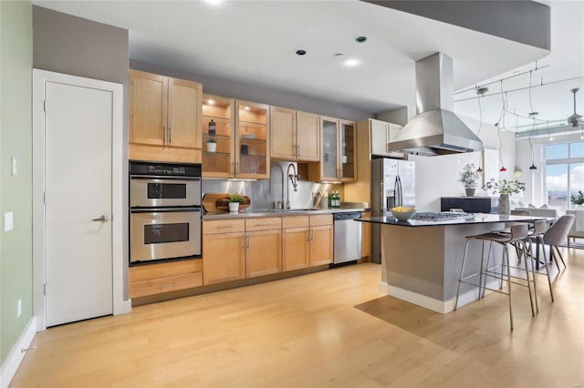 kitchen featuring island exhaust hood, light wood finished floors, light brown cabinetry, appliances with stainless steel finishes, and a sink