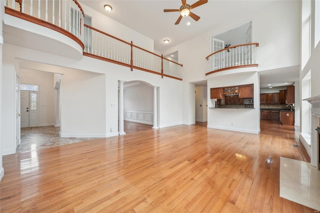 unfurnished living room featuring arched walkways, ornate columns, a ceiling fan, light wood-type flooring, and baseboards