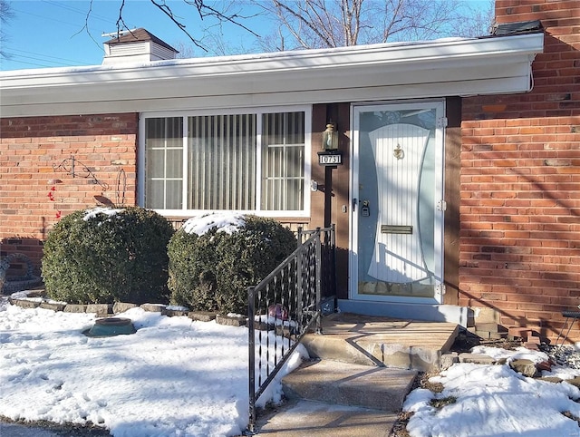 snow covered property entrance featuring a chimney and brick siding