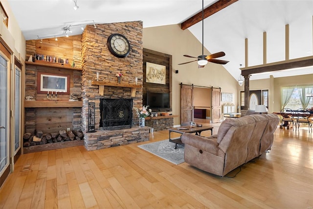 living room featuring ceiling fan, beam ceiling, a stone fireplace, a barn door, and light wood-type flooring