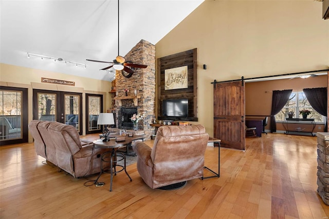 living room with a stone fireplace, high vaulted ceiling, ceiling fan, a barn door, and light wood-type flooring