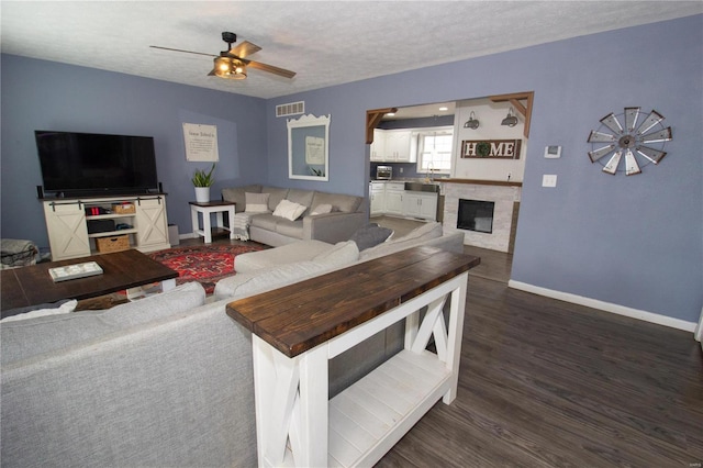 living room featuring a stone fireplace, sink, ceiling fan, dark wood-type flooring, and a textured ceiling