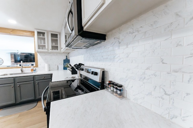 kitchen featuring gray cabinetry, a sink, light countertops, appliances with stainless steel finishes, and glass insert cabinets