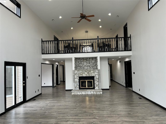 unfurnished living room with a towering ceiling, a ceiling fan, a stone fireplace, wood finished floors, and baseboards