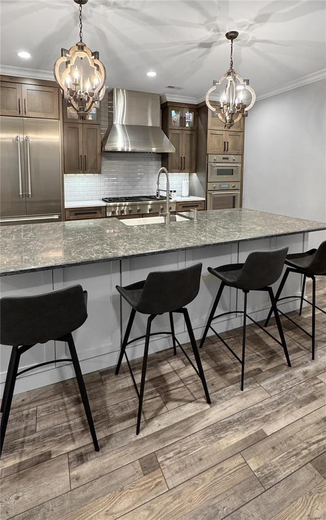 kitchen featuring wall chimney range hood, backsplash, stainless steel appliances, light stone counters, and a breakfast bar area