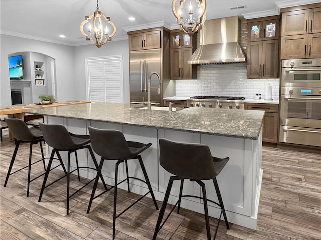 kitchen with appliances with stainless steel finishes, wall chimney exhaust hood, sink, dark hardwood / wood-style floors, and a large island with sink