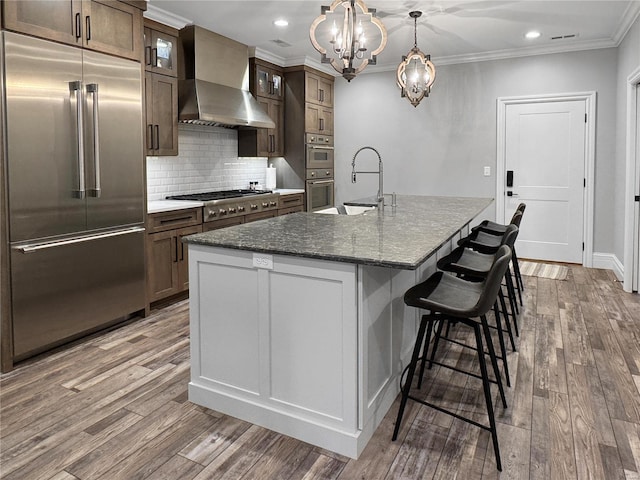 kitchen featuring wall chimney range hood, sink, appliances with stainless steel finishes, dark stone countertops, and a center island with sink