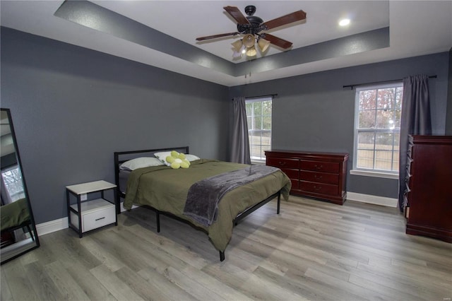 bedroom with ceiling fan, multiple windows, a tray ceiling, and light wood-type flooring