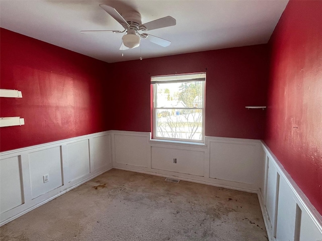 empty room featuring light carpet, a ceiling fan, visible vents, and wainscoting