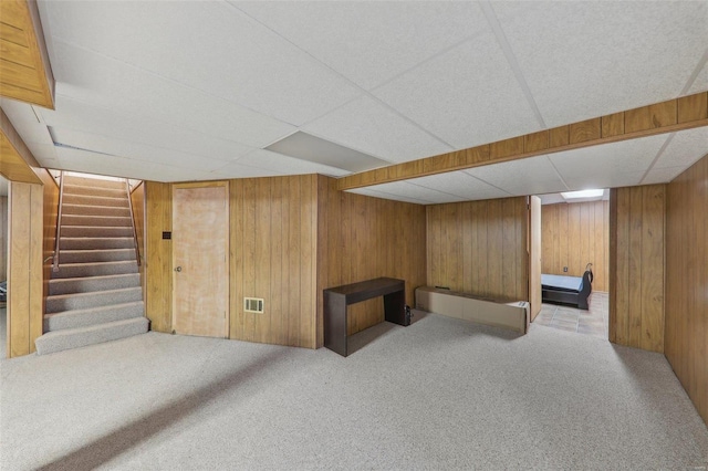 basement featuring light colored carpet, wooden walls, and a drop ceiling