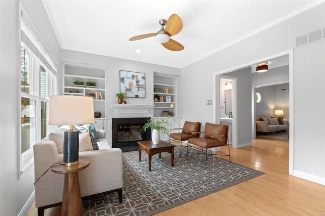 living room with a glass covered fireplace, built in features, a ceiling fan, baseboards, and ornamental molding