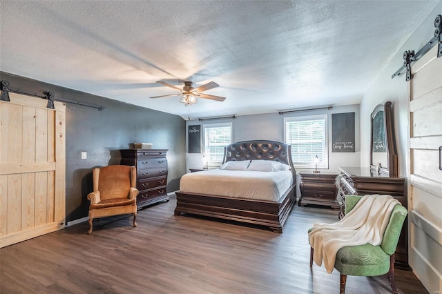 bedroom with ceiling fan, a barn door, dark wood-type flooring, and a textured ceiling