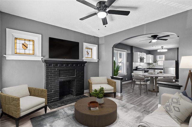 living room featuring a brick fireplace, light wood-type flooring, a textured ceiling, and ceiling fan