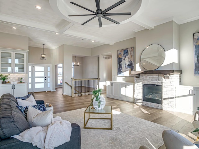 living room featuring a stone fireplace, ceiling fan with notable chandelier, light wood-type flooring, coffered ceiling, and beam ceiling