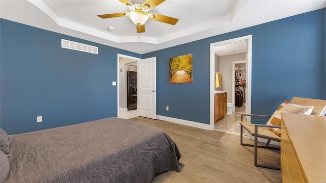 bedroom with black refrigerator, a tray ceiling, ceiling fan, light hardwood / wood-style floors, and ensuite bath