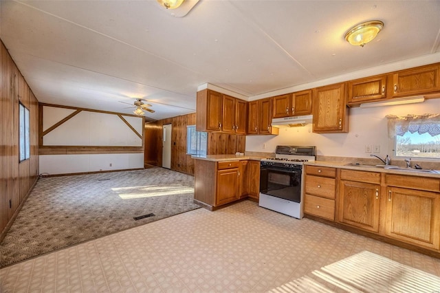 kitchen with white gas stove, wooden walls, light carpet, sink, and ceiling fan