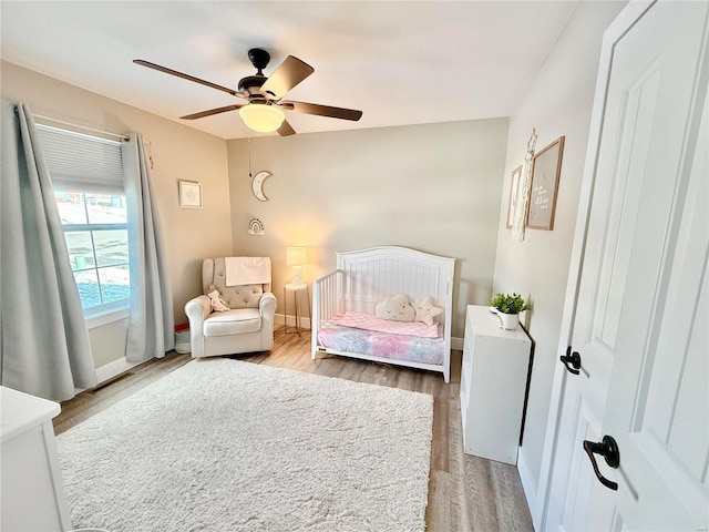 bedroom featuring light wood-type flooring, a nursery area, ceiling fan, and baseboards