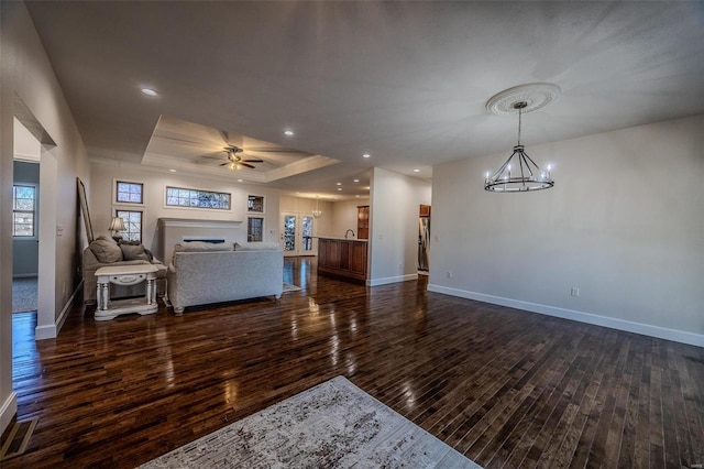 living room with ceiling fan with notable chandelier, dark wood-type flooring, and a raised ceiling