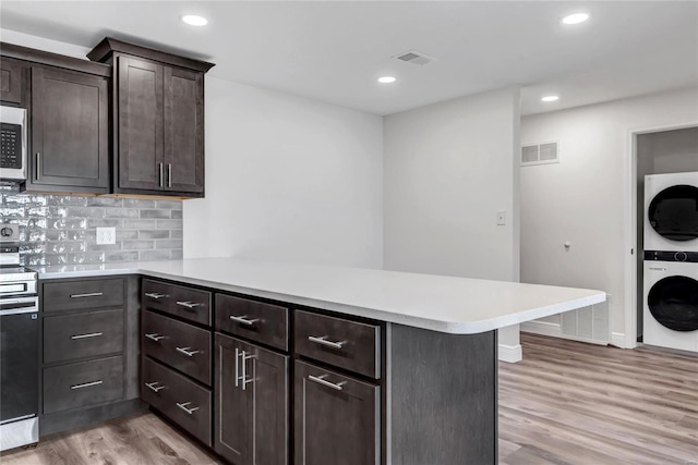 kitchen with dark brown cabinets, appliances with stainless steel finishes, kitchen peninsula, stacked washing maching and dryer, and light hardwood / wood-style floors