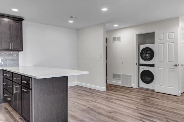 kitchen with backsplash, stacked washer and clothes dryer, kitchen peninsula, dark brown cabinets, and light hardwood / wood-style flooring