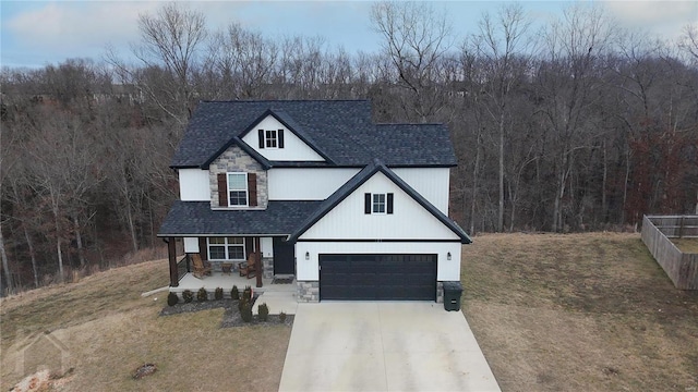 modern farmhouse style home with a shingled roof, concrete driveway, stone siding, a front lawn, and a wooded view