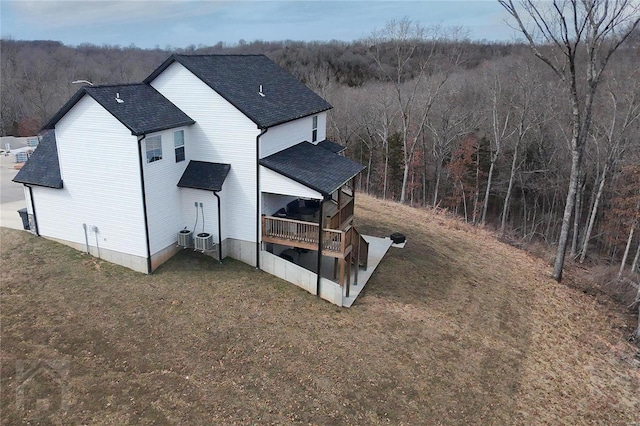 exterior space with a deck, central AC, roof with shingles, a lawn, and a wooded view