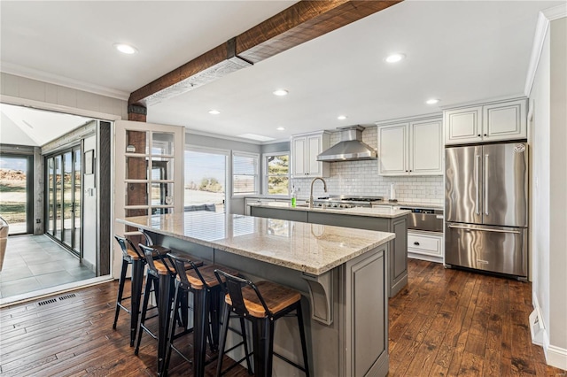 kitchen featuring high end fridge, dark wood finished floors, wall chimney range hood, and an island with sink