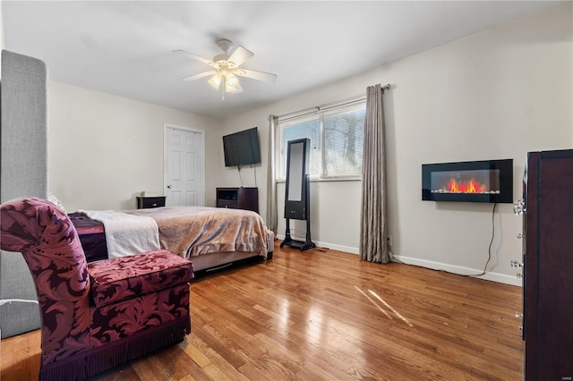 bedroom featuring ceiling fan, a glass covered fireplace, wood finished floors, and baseboards