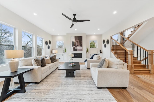 living area featuring a ceiling fan, a glass covered fireplace, recessed lighting, stairway, and wood finished floors