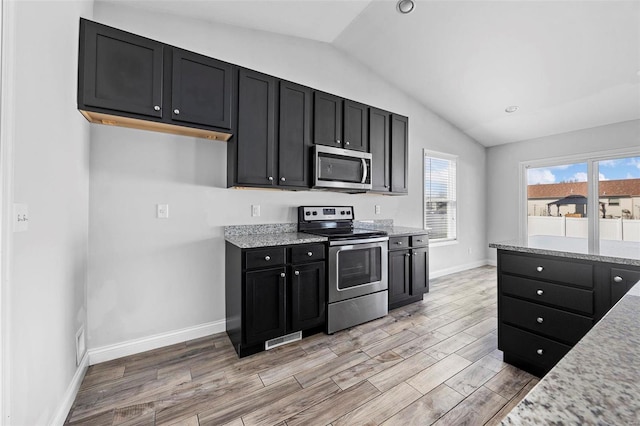 kitchen with lofted ceiling, light stone counters, stainless steel appliances, dark cabinetry, and light wood-type flooring