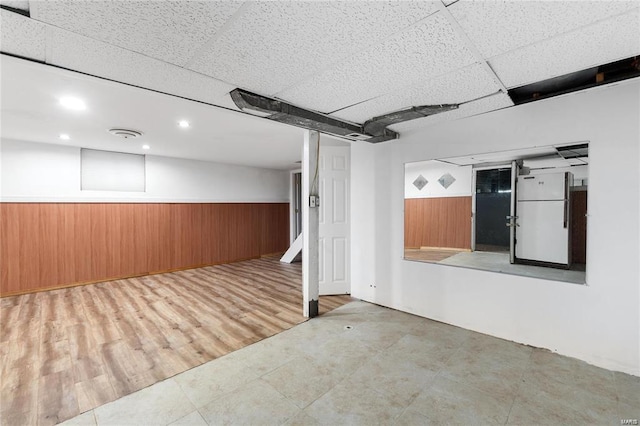 basement featuring white refrigerator, a drop ceiling, and wood walls