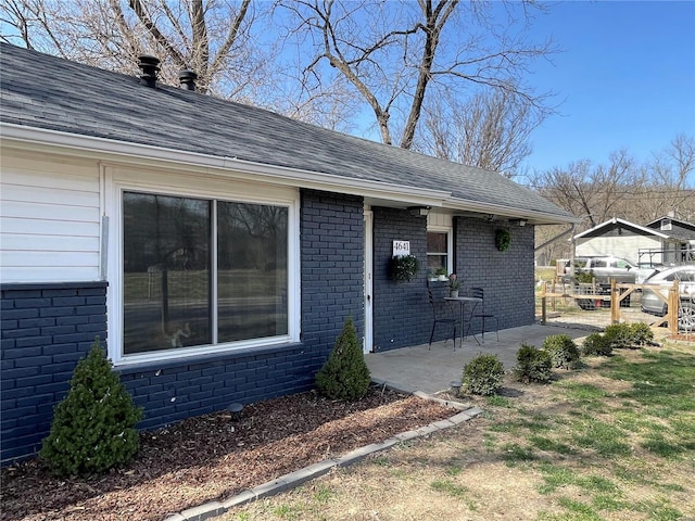 view of side of property featuring brick siding, roof with shingles, and a patio area