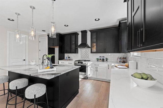 kitchen with pendant lighting, a kitchen island with sink, gas stove, wall chimney range hood, and light wood-type flooring