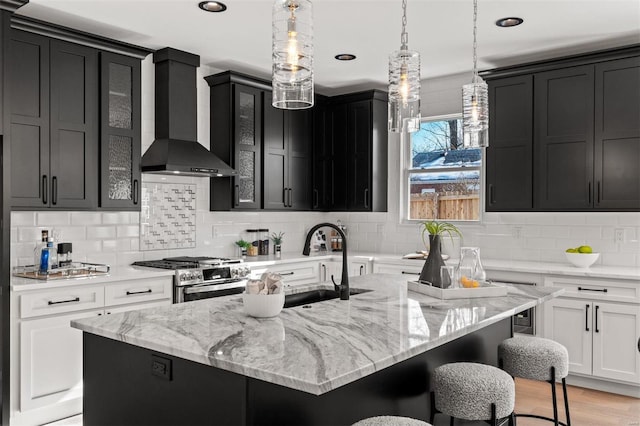 kitchen featuring a kitchen island with sink, white cabinets, stainless steel gas range oven, and wall chimney exhaust hood