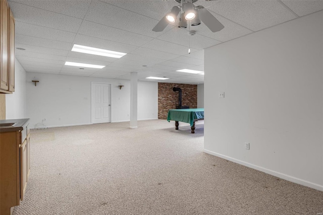 basement with a wood stove, light colored carpet, a drop ceiling, and ceiling fan