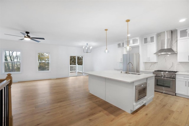 kitchen with hanging light fixtures, appliances with stainless steel finishes, an island with sink, wall chimney range hood, and white cabinets